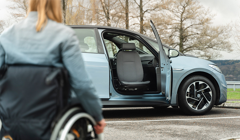 A woman in a wheelchair faces a parked blue car with the passenger door open, the seat is swivelled out of the door opening.